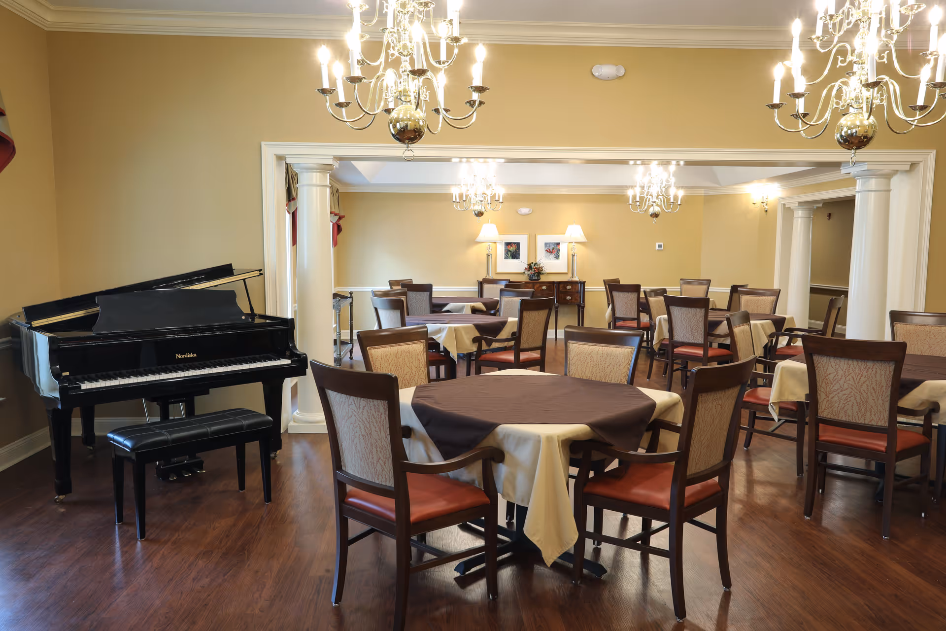 Dining room with round tables and chairs, chandeliers, columns, and a black grand piano.