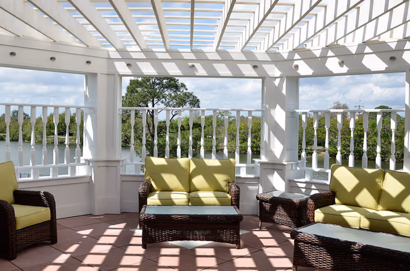 Outdoor seating area with wicker furniture including two sofas and two chairs with light green cushions under a white pergola. The area overlooks a body of water and greenery in the background.