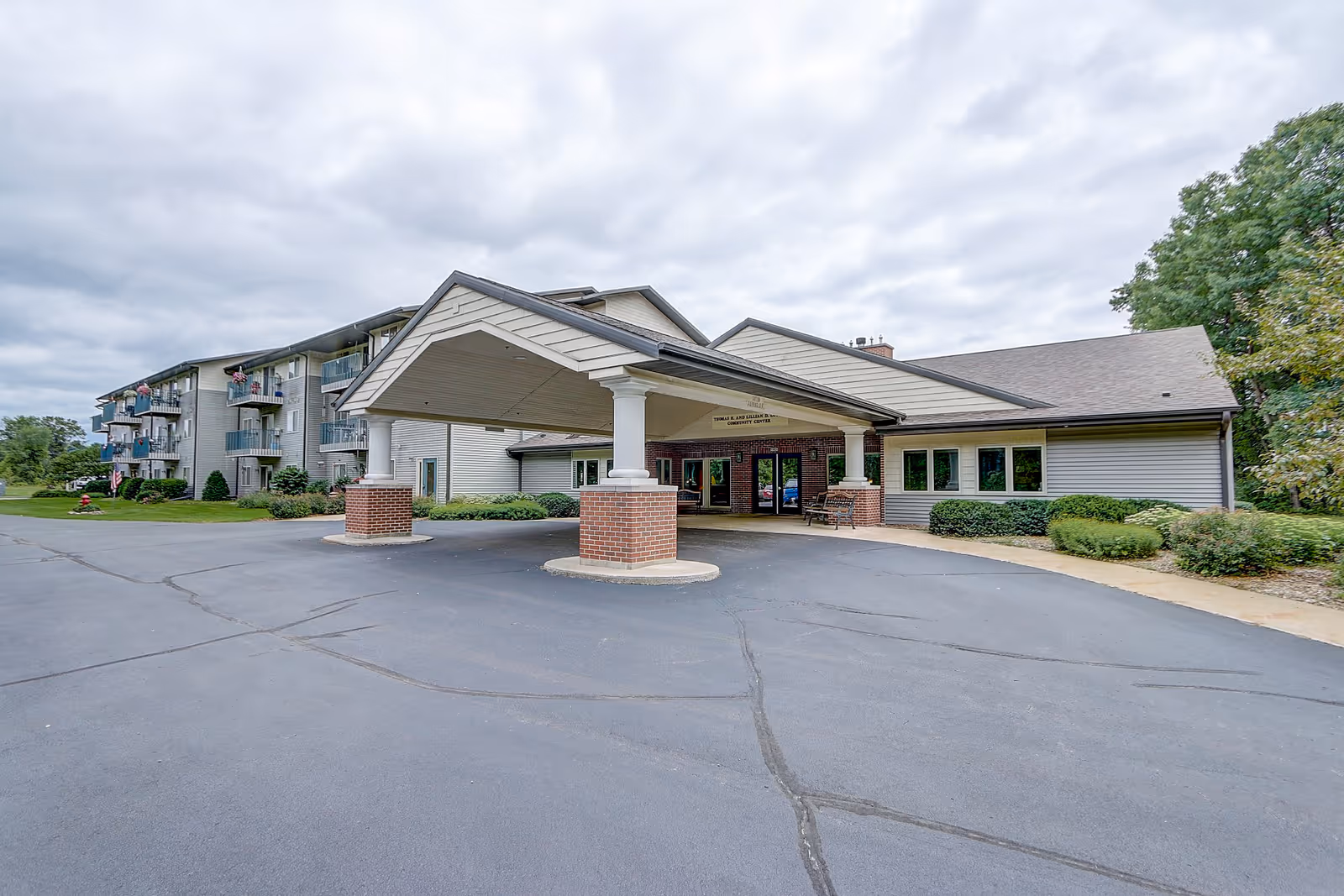 Exterior view of a senior living facility with a covered entrance supported by brick pillars. The building has multiple floors with balconies on the left side and a single-story section on the right. The driveway and parking area are visible in the foreground, with some greenery and trees around the building under a cloudy sky.