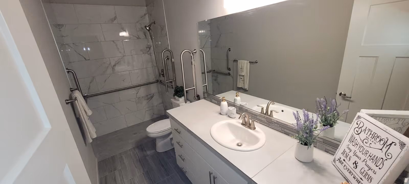 A modern bathroom with a white countertop and sink, a large mirror above the sink, and a toilet next to a walk-in shower with marble-patterned tiles and multiple grab bars. There are towels hanging on the bars, a small potted plant on the counter, and a decorative sign with a message about washing hands.