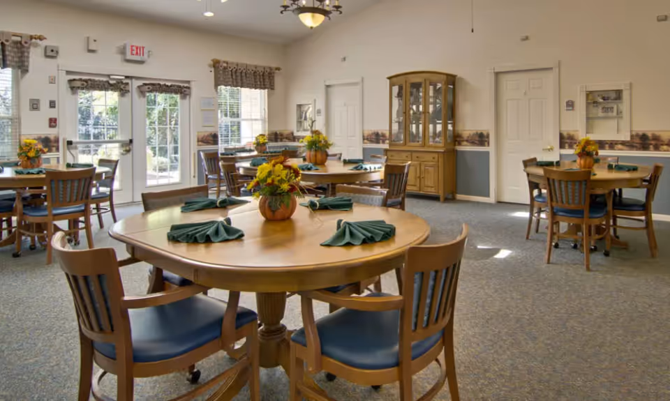 A bright dining room in a senior living facility with round wooden tables set with green folded napkins and floral centerpieces. The room has multiple windows with curtains, a glass door leading outside, and a wooden china cabinet against the wall.