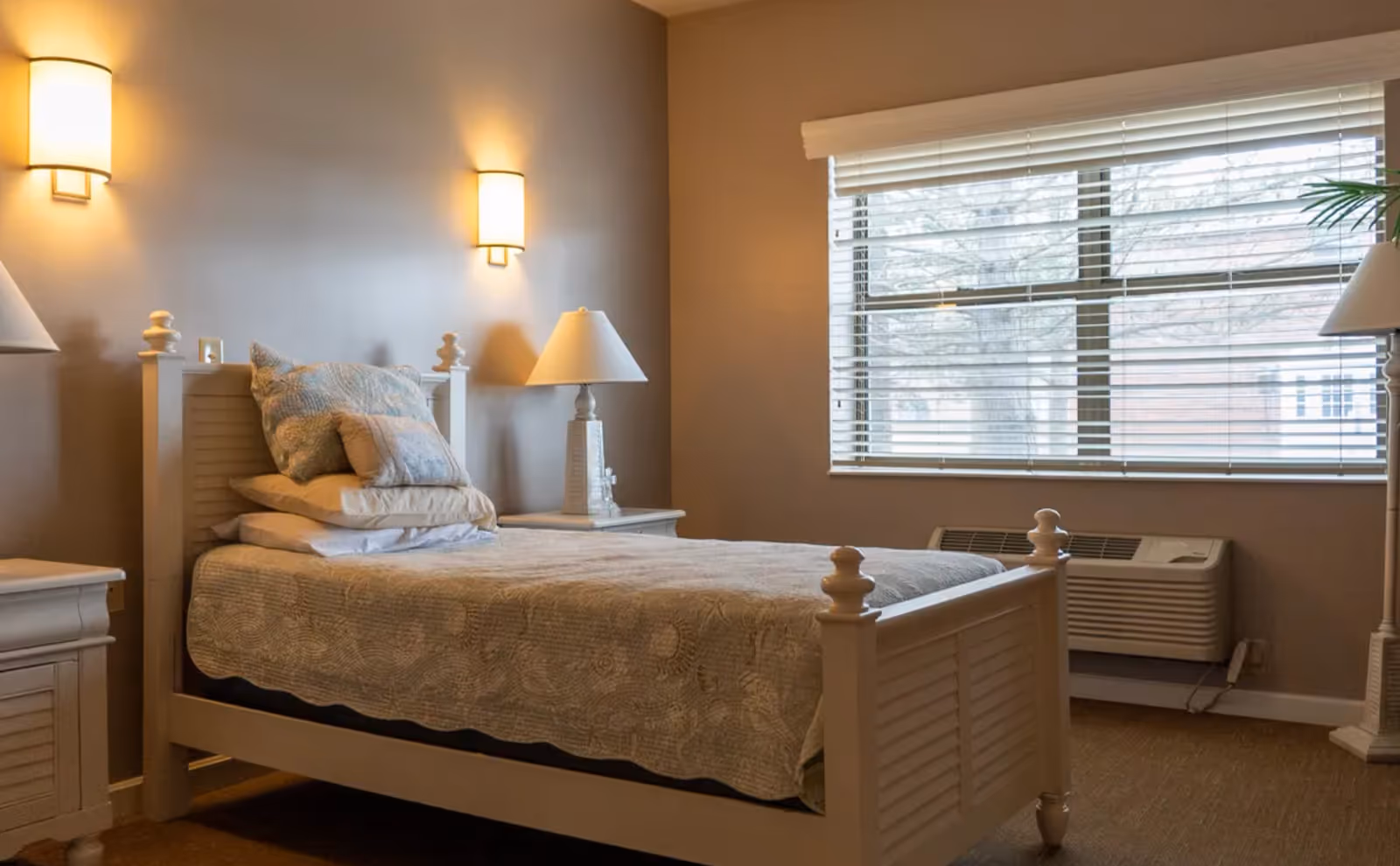 Sunlit furnished bedroom with a single bed, nightstands and lamps beside a large window with blinds.