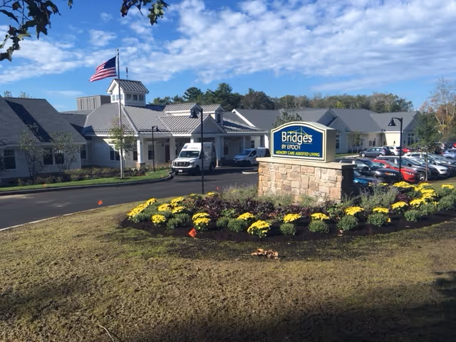 Front exterior of the Bridges by EPOCH senior living facility showing a stone entrance sign, flowerbed, American flag, and parked cars.