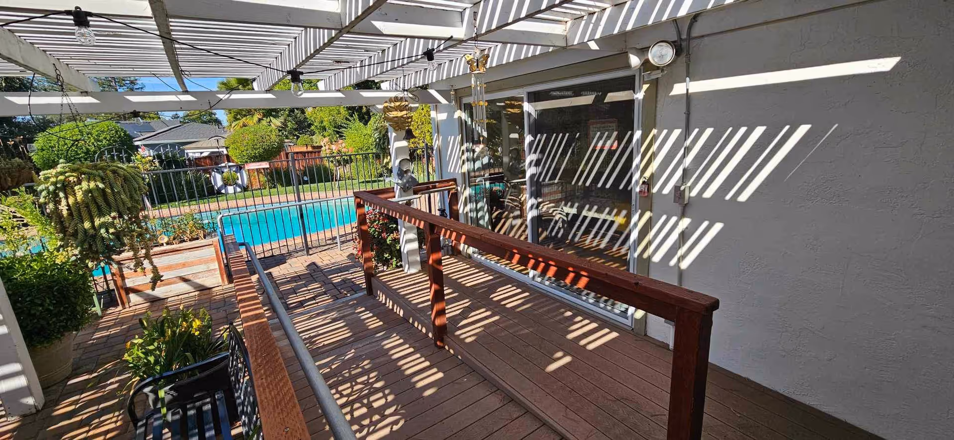 Outdoor patio area with a wooden ramp and railing leading to a fenced swimming pool. The patio has a pergola casting striped shadows on the ground and walls. There are potted plants and greenery around the pool area.