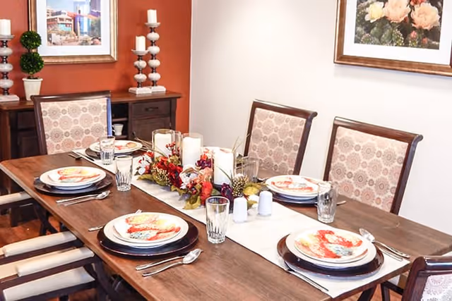 A dining table set for six with plates, glasses, and silverware. The table has a white runner with a centerpiece of candles and autumn-themed decorations. The chairs have patterned upholstery, and the walls feature framed artwork and decorative items.