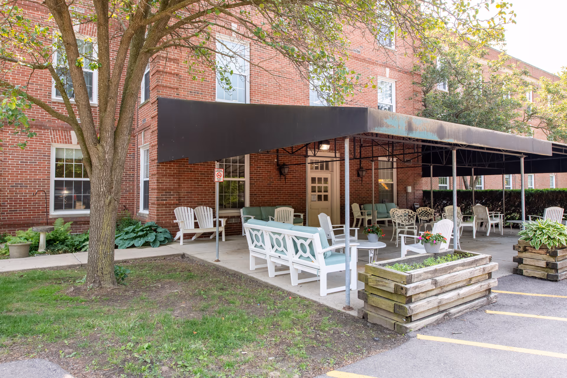 Covered outdoor seating area with white patio furniture and planters in front of a red brick building.