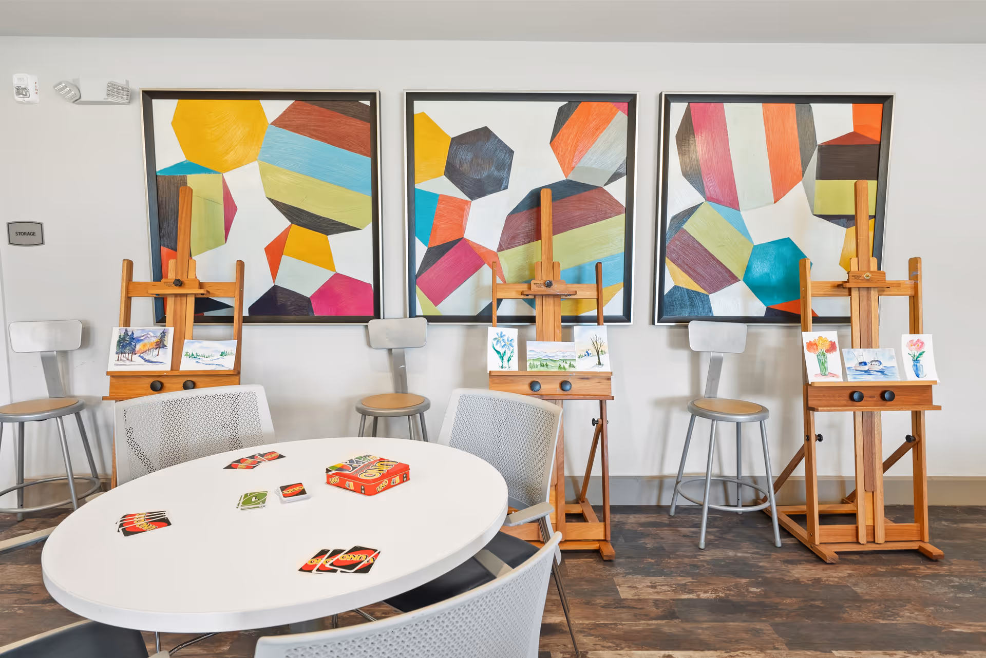 A bright room with a white oval table surrounded by gray chairs. On the table are several Uno cards and a red Uno box. Behind the table are three wooden easels displaying small paintings of nature and flowers. Three colorful abstract paintings hang on the white wall behind the easels. Four metal stools are positioned against the wall.