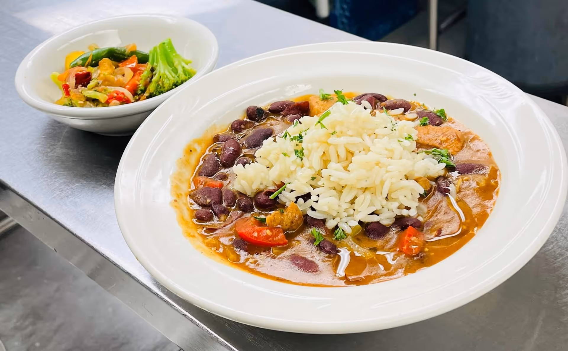 A white bowl filled with a stew containing kidney beans, pieces of meat, and vegetables topped with white rice, accompanied by a smaller bowl of mixed steamed vegetables including broccoli, carrots, and green beans, placed on a metal surface.