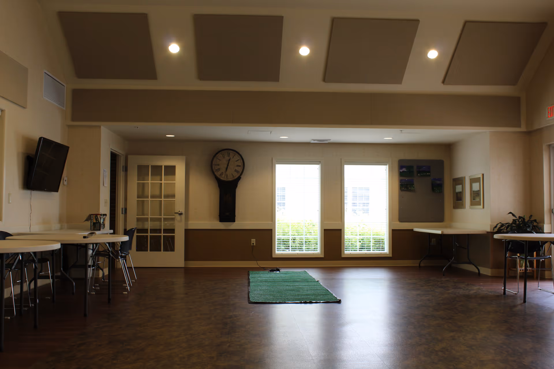 Spacious multipurpose common room with folding tables and chairs, a wall clock, two windows, and a small green rug on the floor.