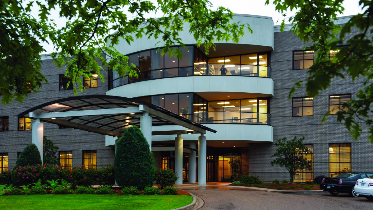 Exterior view of a multi-story brick building with large windows and a covered entrance. The building is surrounded by well-maintained landscaping including bushes, trees, and a green lawn. Several cars are parked near the entrance.
