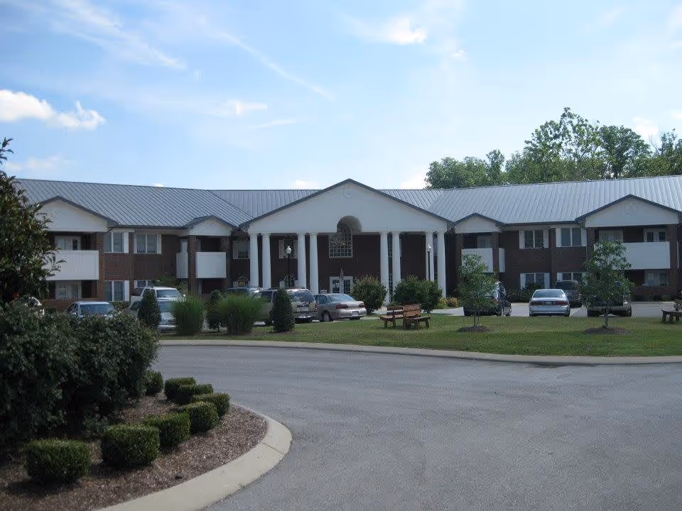 Front exterior view of a two-story senior living facility building with a metal roof, white columns at the entrance, parked cars, green lawn, and small trees and bushes under a partly cloudy sky.