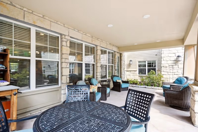 Covered outdoor patio area with stone walls and multiple windows. The space is furnished with black metal tables and chairs with blue cushions, as well as wicker armchairs with blue cushions and decorative pillows. There are plants and wall-mounted lights on the far wall.