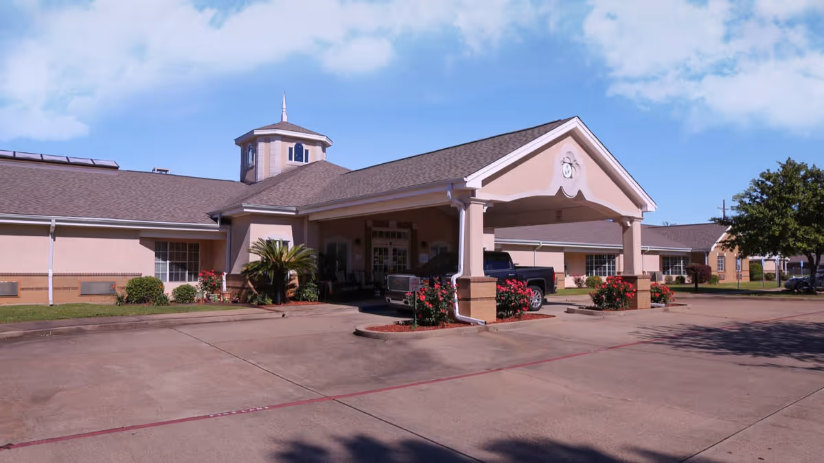 Exterior view of a single-story senior living facility building with a covered entrance, landscaped bushes, and a parked pickup truck under the entrance canopy. The sky is clear with some clouds.