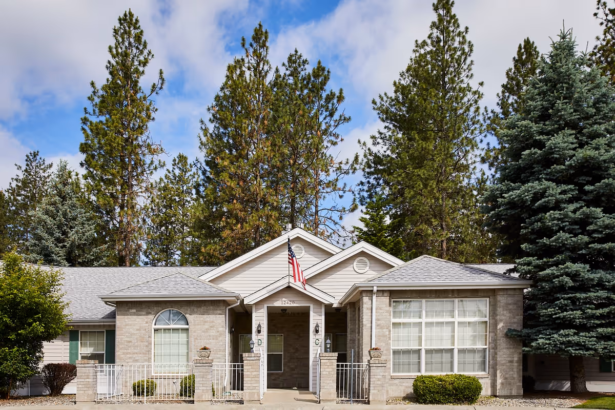 Single-story brick building front with an American flag over the entry, large windows, and tall pine trees in the background.