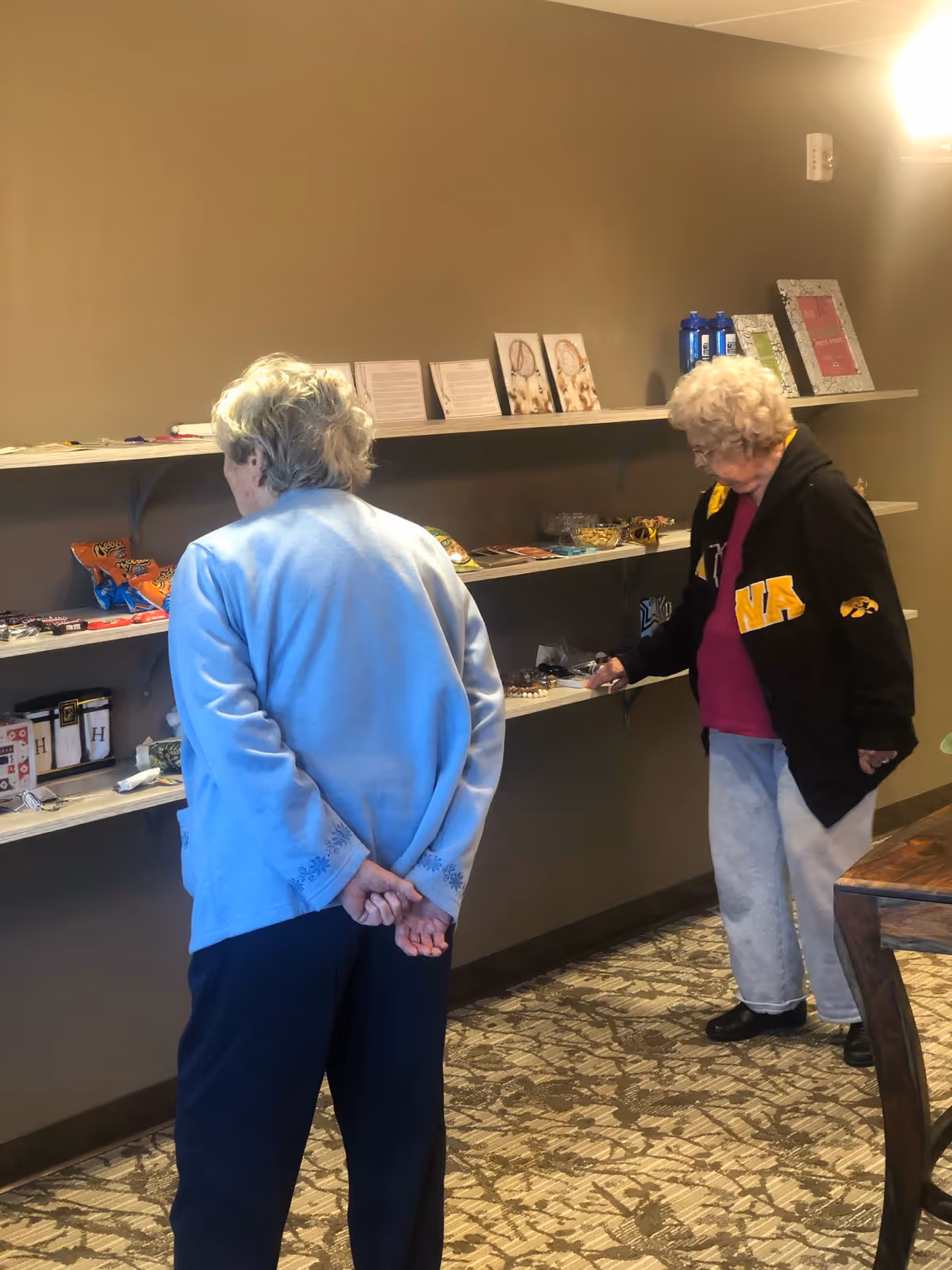 Two elderly women standing in front of wall-mounted shelves with various snacks, small items, and framed pictures in a room with patterned carpet and beige walls.
