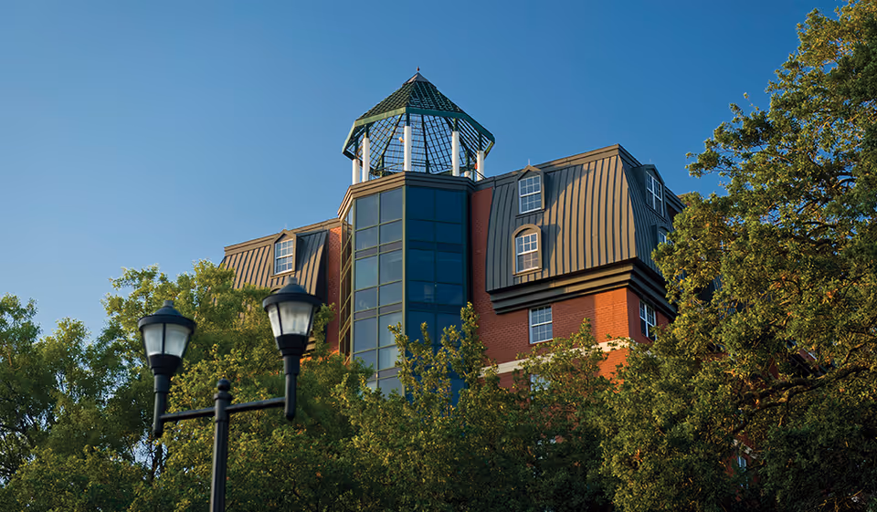 Exterior view of a multi-story brick building with a distinctive glass and metal tower structure on the roof, surrounded by green trees under a clear blue sky. Two black street lamps are visible in the foreground.