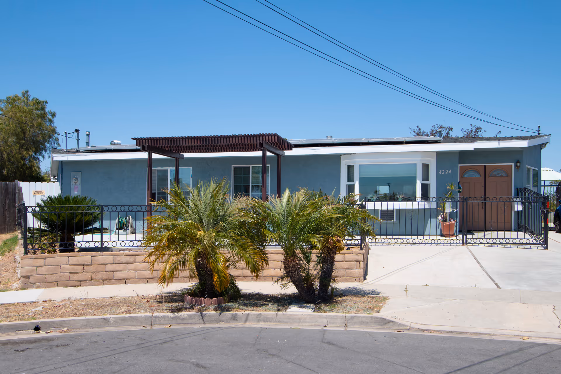 Single-story blue residential building with a small front yard featuring palm plants, a black metal fence, and a concrete driveway. The building has a pergola over the front patio and two brown front doors under a clear blue sky.