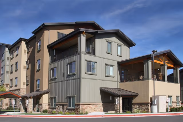 Exterior view of a multi-story senior living facility building with a combination of beige and gray siding, stone accents at the base, multiple windows, and covered balconies under a clear blue sky.