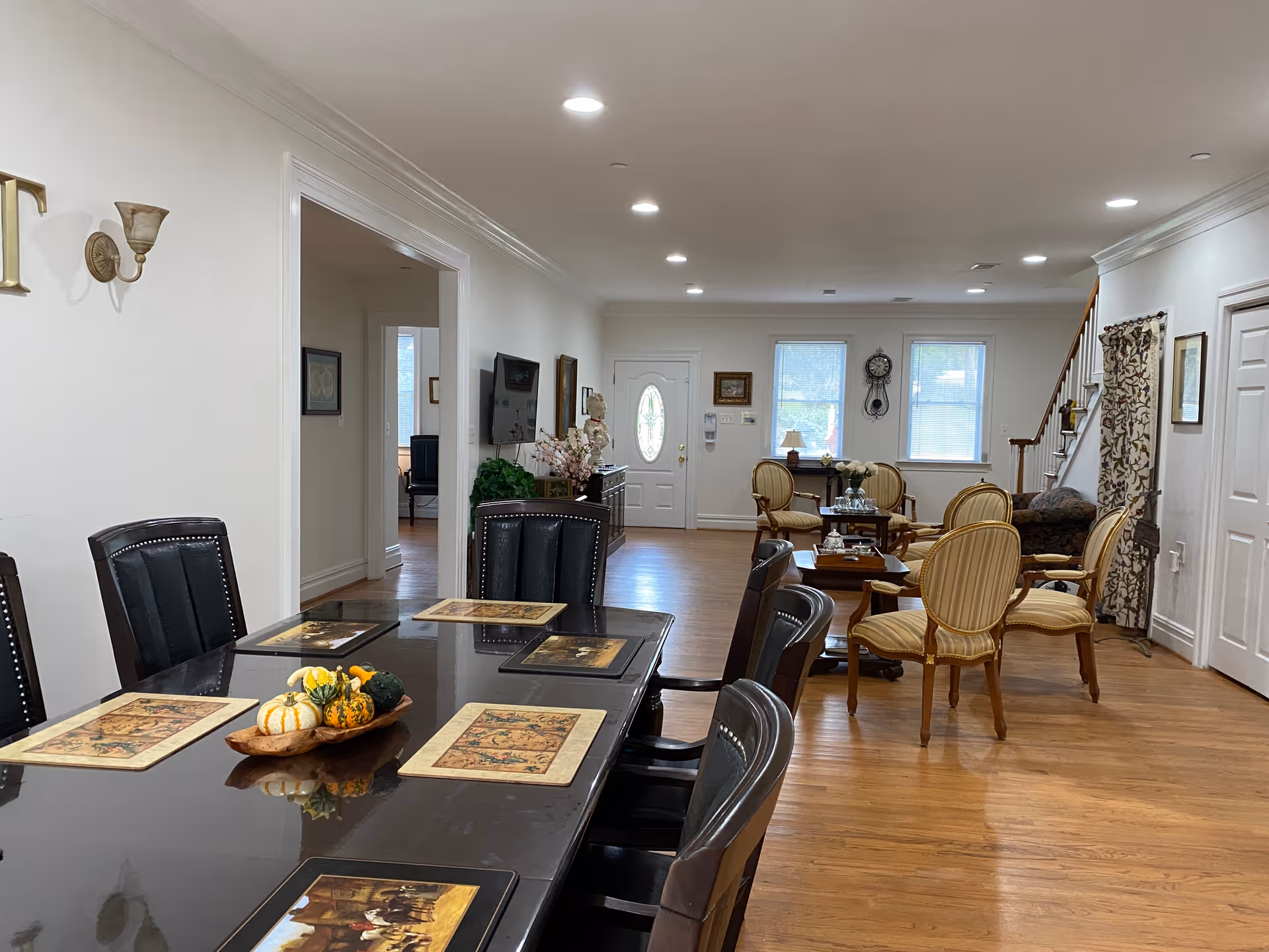 Interior view of a spacious assisted living facility common area with a large dark dining table set with six black chairs and decorative placemats. The table has a centerpiece with small decorative gourds. In the background, there is a sitting area with several upholstered chairs arranged around small tables. The room has wooden flooring, white walls, recessed ceiling lights, and windows letting in natural light. A staircase is visible on the right side of the room.