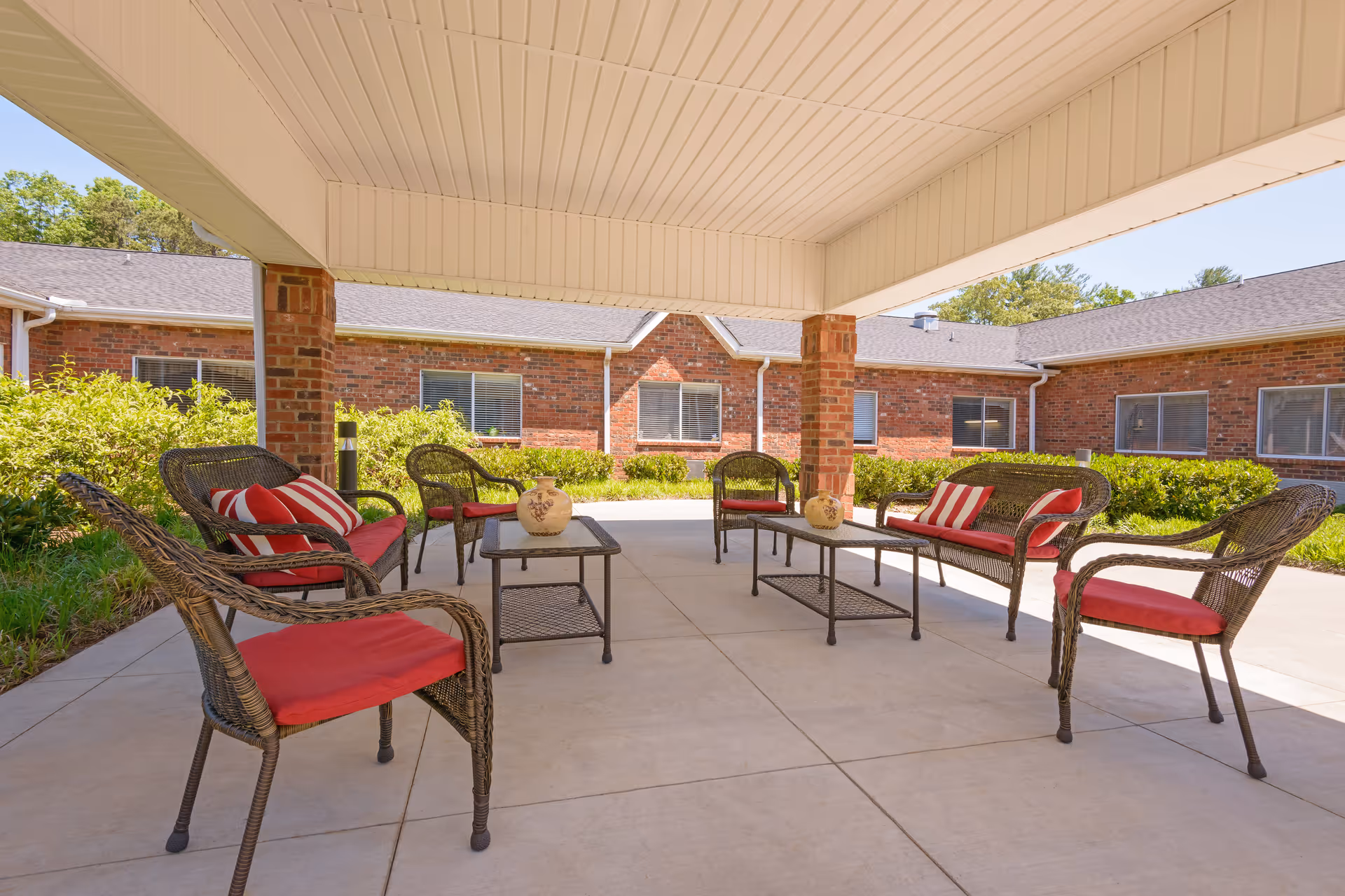 Covered outdoor patio area with wicker chairs and sofas featuring red cushions and striped pillows, two coffee tables with decorative vases, surrounded by a brick building and greenery under a clear blue sky.