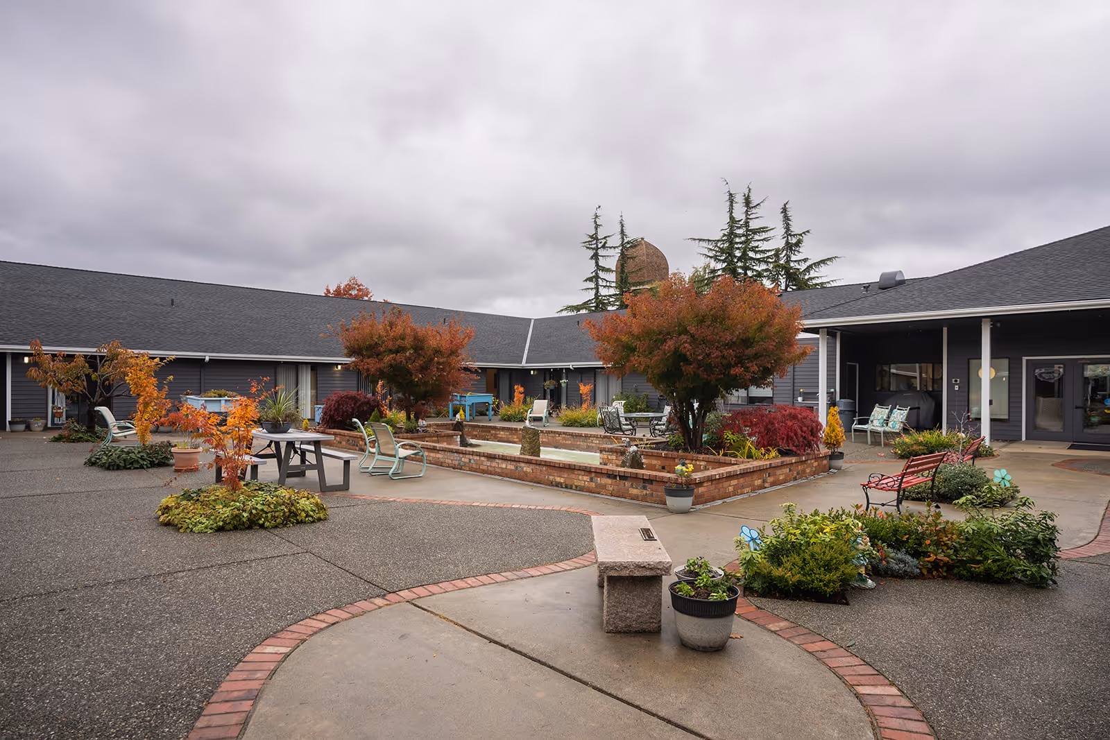 Outdoor courtyard with brick raised planters, seating, and single-story buildings under a cloudy sky.