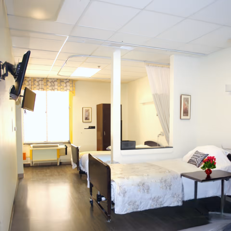 A bright and clean room with two hospital-style beds separated by a partial wall with a curtain. Each bed has white bedding and pillows, with a small table next to one bed holding a vase of red flowers. A flat-screen TV is mounted on the wall, and a window with patterned curtains lets in natural light. The room has light-colored walls and dark flooring.