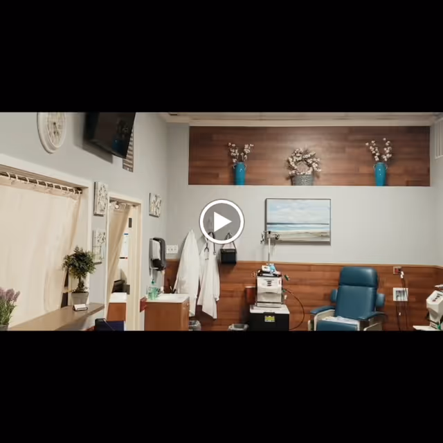 Interior of a medical or nursing facility room with a blue recliner chair, medical equipment, a sink with hand sanitizer, white coats hanging on hooks, decorative vases with flowers on a wooden shelf, and a landscape painting on the wall.