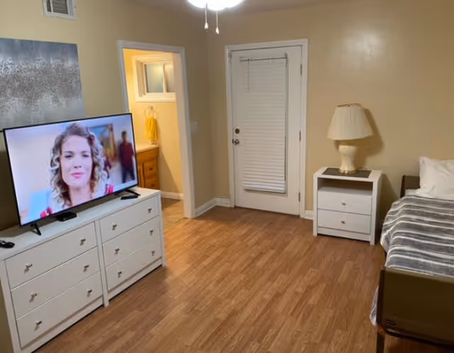 A senior living facility bedroom with wooden flooring, a bed with a gray striped blanket, a white nightstand with a lamp, a white dresser with a flat-screen TV showing a woman, and a door with a window blind. There is an open doorway leading to a bathroom with a yellow towel visible.