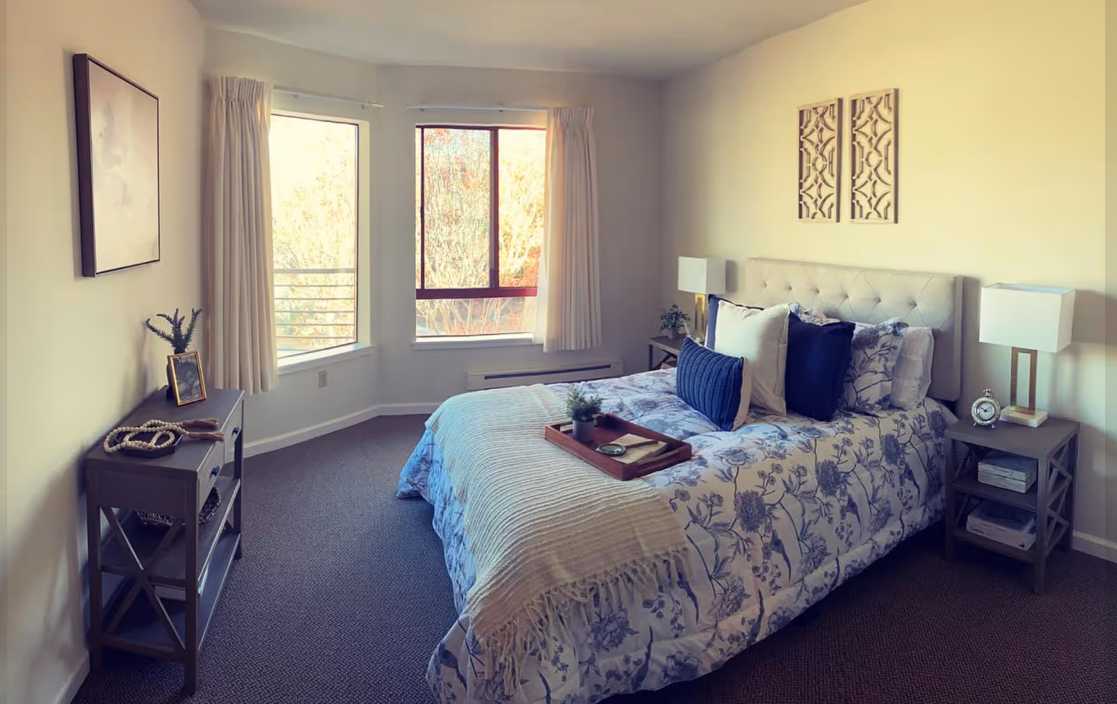 A cozy bedroom with a bed dressed in floral patterned bedding and multiple pillows. The bed has a tufted headboard and a wooden tray with decorative items on top. There are two nightstands on either side of the bed, each with a modern lamp and small decor. The room has two large windows with cream-colored curtains, allowing natural light to brighten the space. A small console table with decor and a framed picture is placed against the opposite wall.