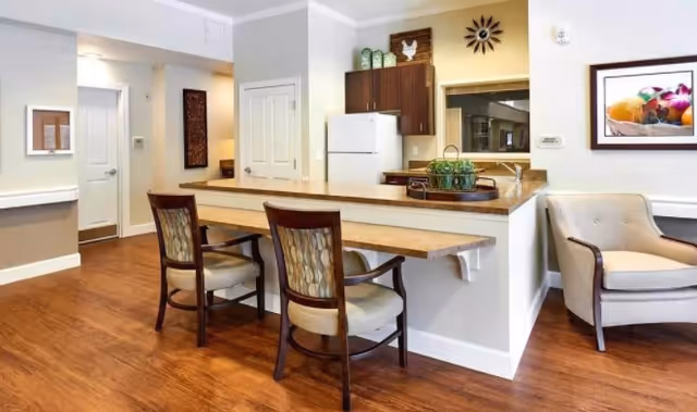 Interior view of a memory care facility showing a kitchen area with a refrigerator, cabinets, and a counter with two chairs. There is a beige armchair to the right and framed artwork on the wall. The floor is wooden and the walls are painted light gray.