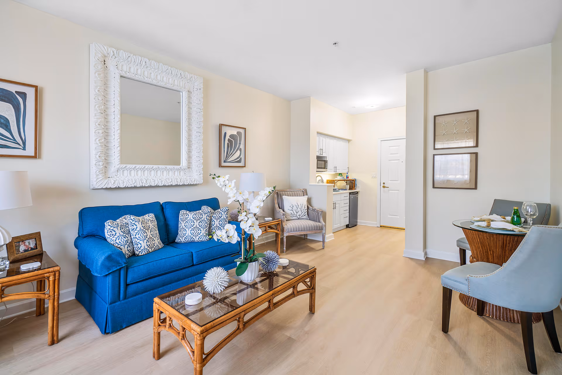 Bright furnished living room with a blue sofa, glass-top coffee table, small dining nook and a kitchenette in the background.
