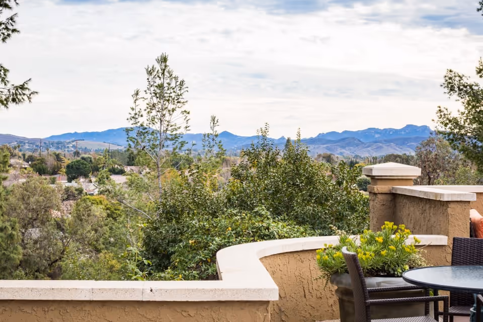 Outdoor patio area with a round glass table and wicker chairs, surrounded by a beige stucco wall. Green shrubs and trees are visible beyond the patio, with mountains in the distance under a partly cloudy sky.