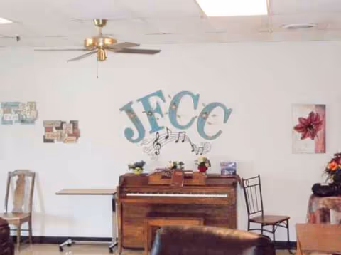Piano against wall with JFCC initials, floral decor, and seating in a lounge.