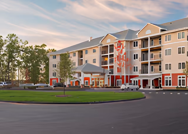 Exterior view of a multi-story senior living facility building with a covered entrance, surrounded by a circular driveway and green lawn, under a partly cloudy sky at sunset.
