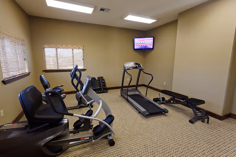 A small fitness room with exercise equipment including two recumbent stationary bikes, a treadmill, a weight bench, and a rack of dumbbells. The room has beige walls, carpeted floor, two windows with woven blinds, and a wall-mounted TV in the corner.