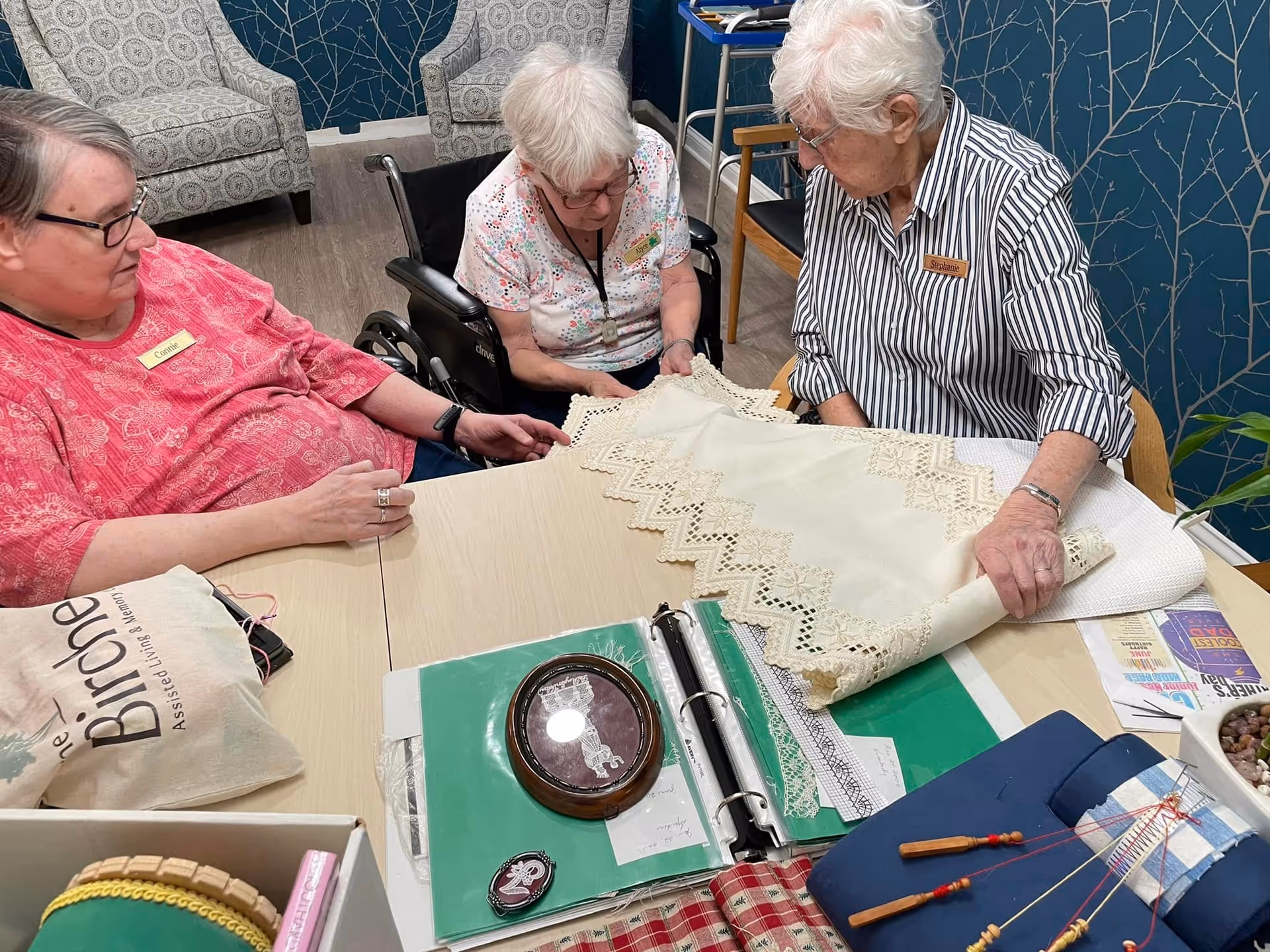Three elderly women sitting around a table engaged in a craft activity involving lace and fabric. One woman is in a wheelchair, and the others are seated on chairs. The table has various craft materials, including a binder with lace samples, bobbins, and a bag labeled 'Birches Assisted Living and Memory Care.' The room has patterned armchairs and a blue wall with a tree branch design.