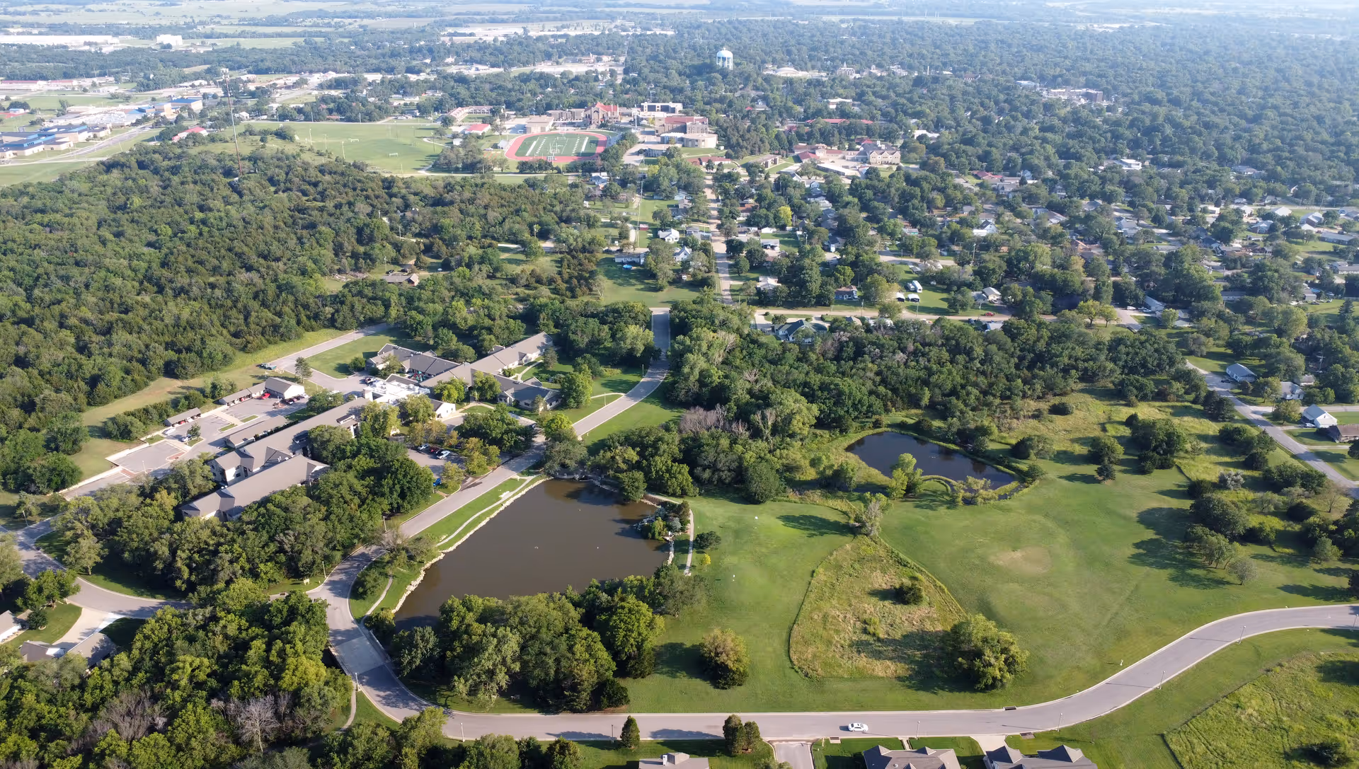 Aerial view of a suburban area with a mix of residential buildings, green spaces, and two small ponds. Roads wind through the landscape, and there is a cluster of buildings surrounded by trees on the left side. The background shows more houses and a distant water tower.