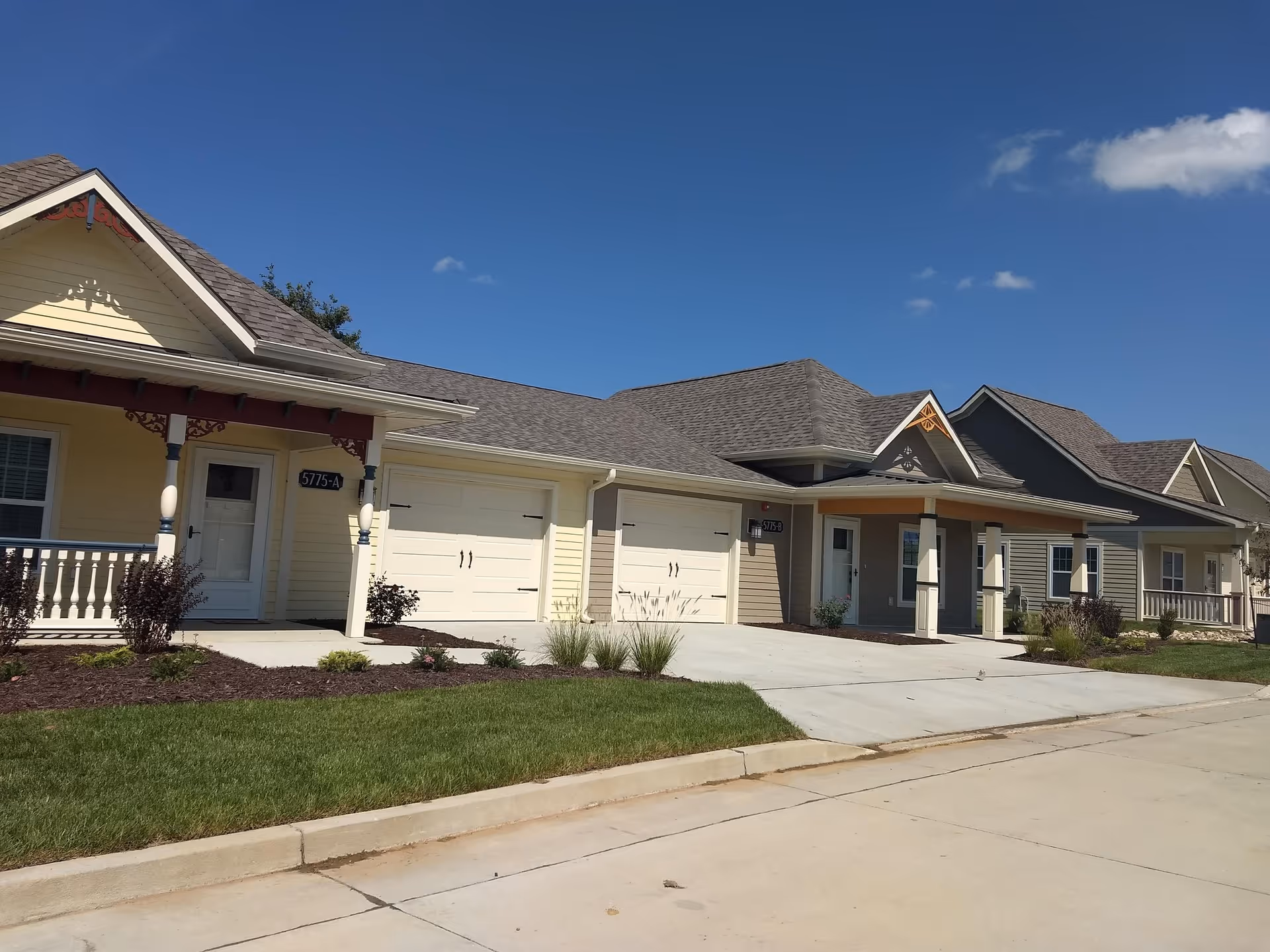 Exterior view of a row of single-story residential buildings with garages, porches, and well-maintained lawns under a clear blue sky.