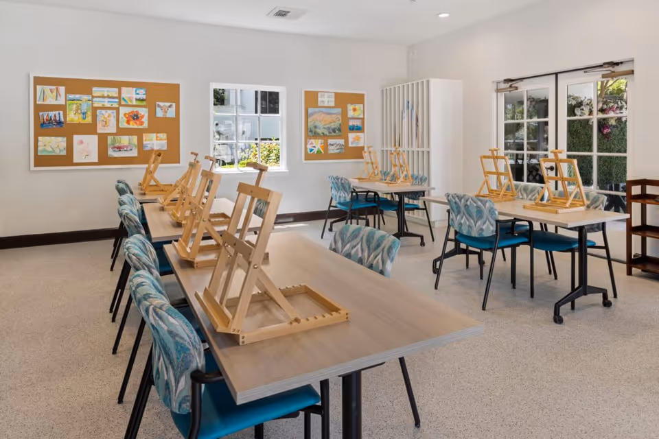 A bright, spacious art room with several tables and chairs arranged in rows. Each table has wooden easels set up, and colorful artwork is displayed on two cork bulletin boards on the walls. Large windows and glass doors let in natural light, and the room has a clean, organized appearance.