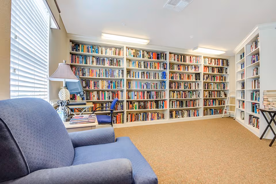 A cozy library room with white built-in bookshelves filled with books along two walls. There is a blue upholstered armchair in the foreground, a small table with a lamp and a computer monitor near a window with white blinds, and a small stack of books on a folding table on the right side.