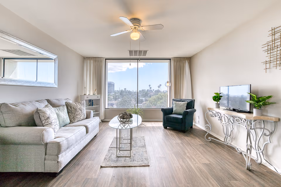 Bright living room with a sofa, armchair, glass coffee table, TV on a decorative console, and a large window with a city view.