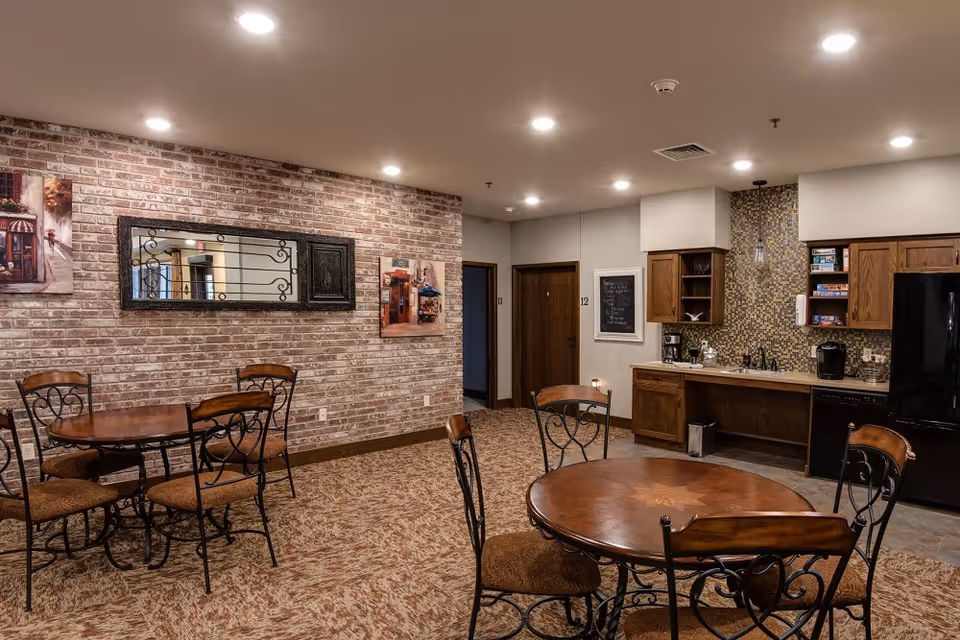 Communal dining area with round wooden tables and chairs, a kitchenette, and a brick accent wall.