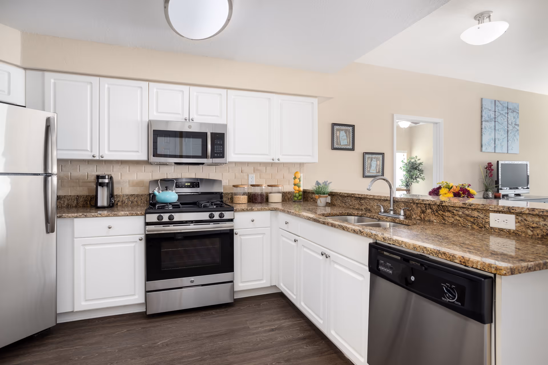 Modern kitchen with white cabinets, stainless steel refrigerator, stove, microwave, and dishwasher. The countertop is granite with a sink and faucet, decorated with a small plant, a container of lemons, and a flower arrangement. The kitchen opens to a living area with a TV and wall art.