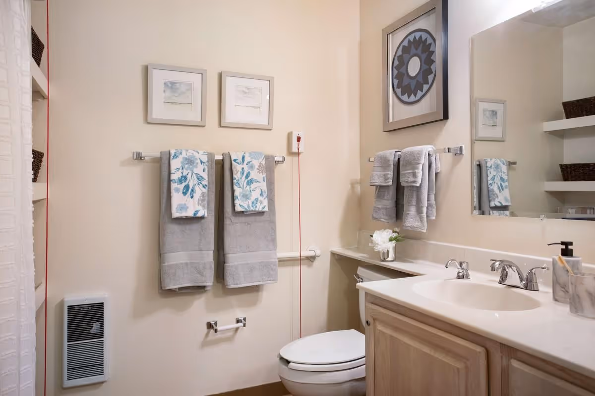 Clean accessible bathroom with a sink and vanity, toilet, towel racks with gray and patterned towels, a wall mirror, and an emergency pull cord.