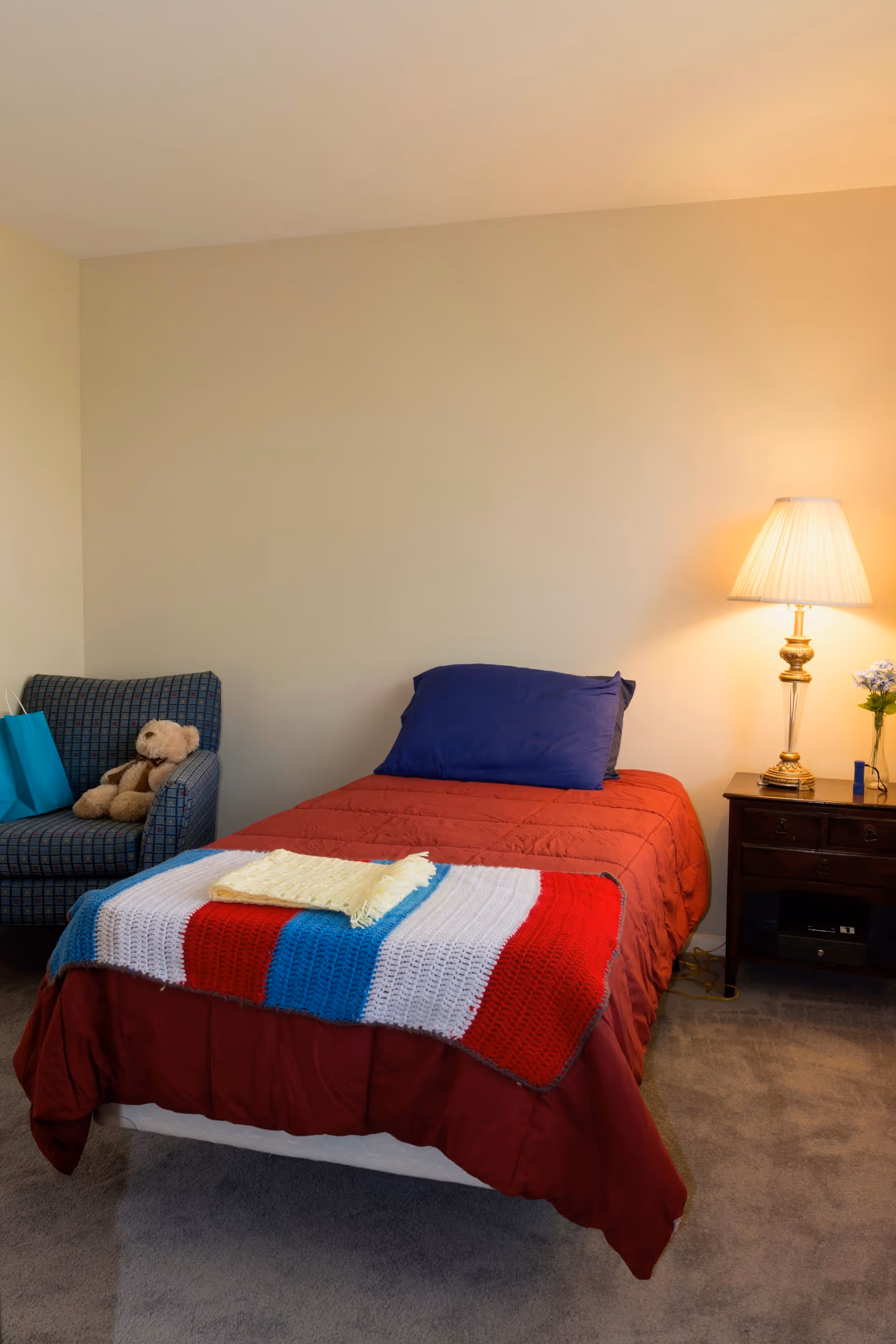 A cozy bedroom with a single bed covered in a red comforter and a colorful knitted blanket at the foot. A blue pillow rests on the bed. To the left, there is a patterned armchair with a teddy bear and a blue gift bag on it. On the right side, a wooden nightstand holds a classic lamp with a pleated shade and a small vase with flowers. The room has beige walls and carpeted flooring.