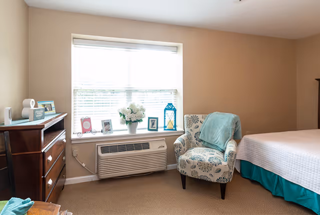 A cozy bedroom with a large window covered by white blinds. Below the window is an air conditioning unit. On the window sill are decorative items including framed photos, a white flower pot with flowers, and a blue lantern. To the right of the window is a patterned armchair with a teal throw draped over it. Part of a bed with a white quilt and teal bed skirt is visible on the right. On the left side of the image is a wooden dresser with drawers and some small decorative items on top.