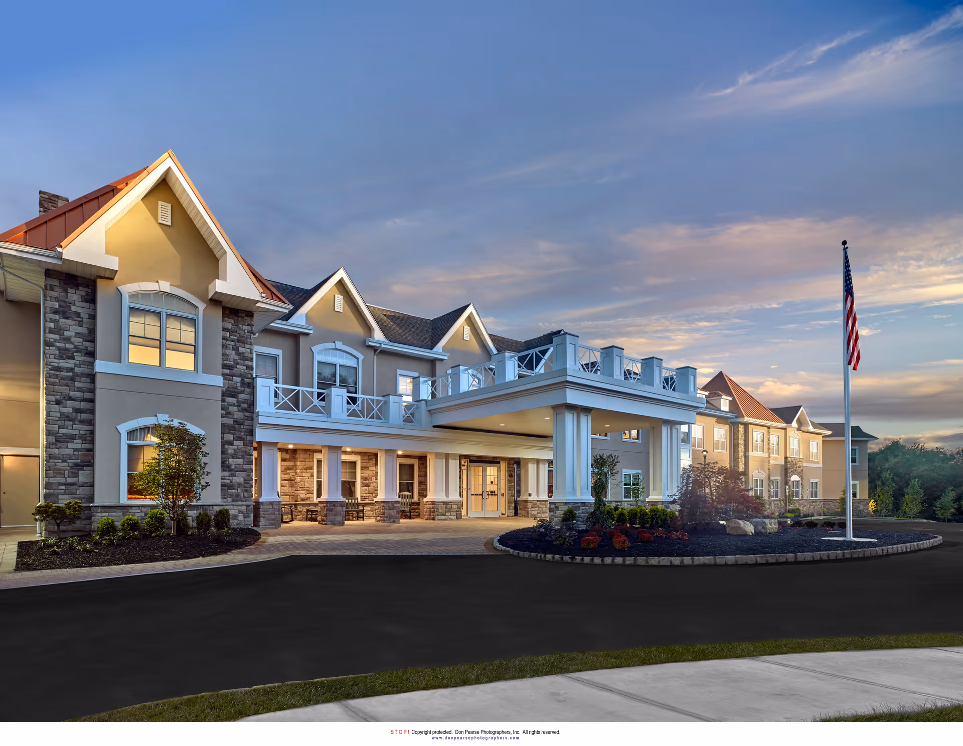 Exterior front view of a senior living facility building with beige walls, stone accents, white trim, and a covered entrance. The building has multiple windows and a balcony area above the entrance. There is a landscaped circular driveway with plants and an American flag on a flagpole. The sky is partly cloudy during sunset.