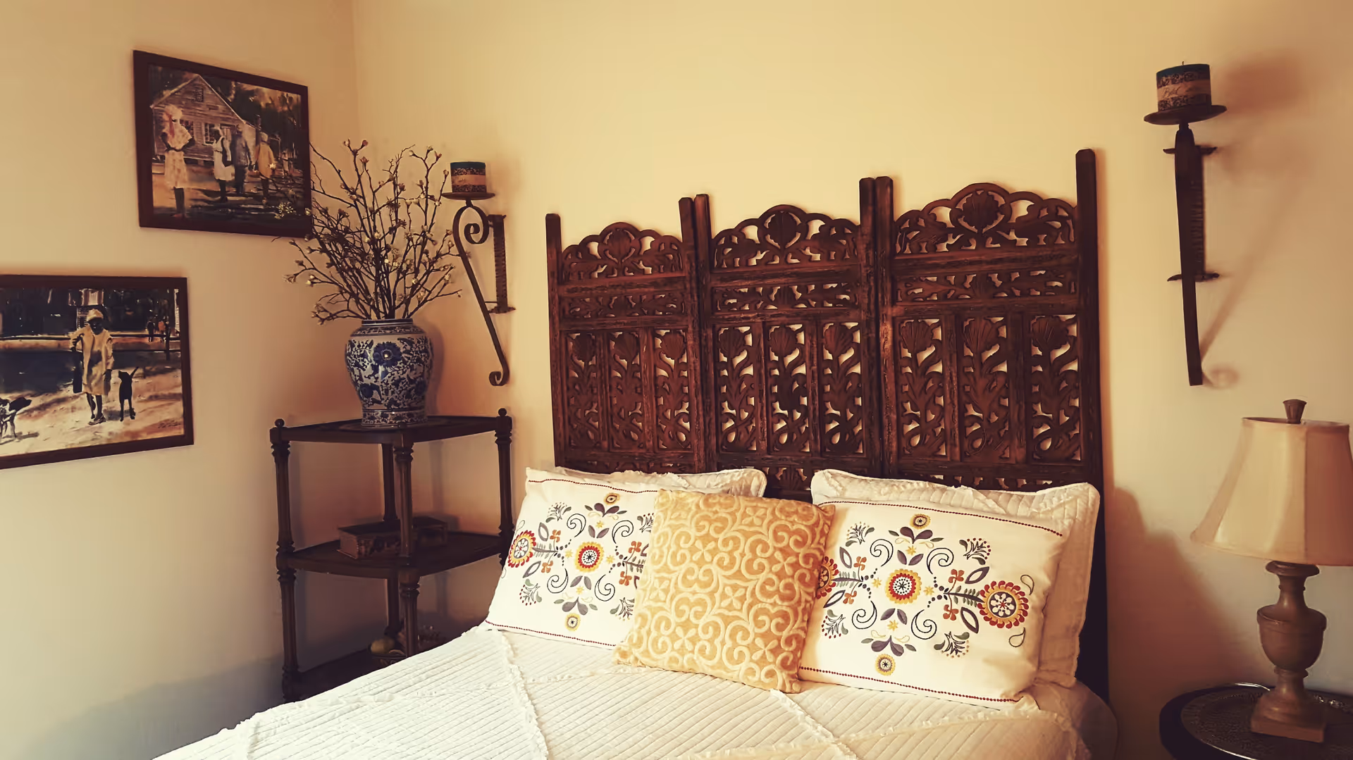 A cozy bedroom with a carved wooden headboard, a bed with white bedding and decorative pillows featuring floral patterns. On the left side, there is a wooden corner shelf with a blue and white vase holding branches. Two framed paintings hang on the wall above the shelf. On the right side, there is a small wooden table with a lamp and two wall-mounted candle holders with candles.