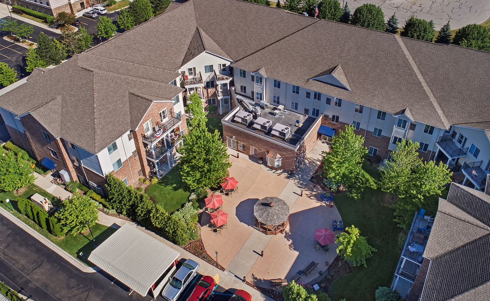 Aerial view of American House Sterling Woods senior living facility showing a large multi-story building surrounding a central courtyard with a gazebo, red umbrellas, outdoor seating, and green trees. Cars are parked in a lot adjacent to the building.