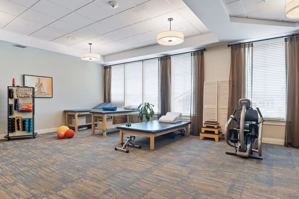 Well-lit therapy/exercise room with treatment tables, exercise equipment, therapy balls, and large windows with blinds.